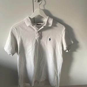 Vit pikétröja från Ralph Lauren - Klassisk vit pikétröja från Ralph Lauren med korta ärmar och broderad marinblå logga på bröstet. Tröjan har krage och knäppning framtill. Perfekt för en clean och stilren look.