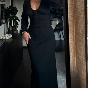 Svart långklänning från NA-KD - Supersnygg svart långklänning från NA-KD med ribbad struktur och v-ringning. Klänningen har långa ärmar och snörning framtill som ger en cool detalj. Figurnära passform som framhäver siluetten. 