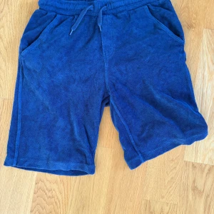 Blå frottéshorts från Lindex 158-164 - Mysiga marinblå shorts från Lindex i frottématerial, perfekta för chill dagar. De har snörning i midjan, två sidofickor och en bakficka. Sköna att dra på sig efter badet eller när du bara vill vara bekväm. Köpta för ca 500 kr och mitt pris är 149kr 