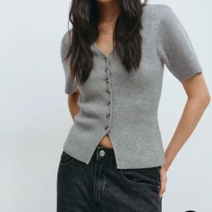 Grå kortärmad stickad topp - Snygg grå stickad topp med v-ringning och korta ärmar. Toppen har knappar framtill och en figurnära passform som ger en clean och stilren look. Perfekt att matcha med jeans eller kjol för en chill men ändå uppklädd vibe. Använd 1 gång så nästan helt ny!