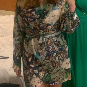 Blommig omlottklänning från Zara - Säljer en färgstark kort omlottklänning från Zara med blommigt mönster i grönt, blått, beige och inslag av orange och gult. Klänningen har lång ärm, v-ringning och knyt i midjan för en snygg siluett. Använd 1 gång💕☺️