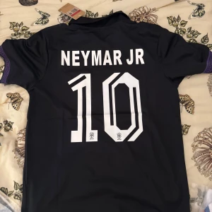Svart Brasil Neymar JR 10 Nike tröja - Säljer en svart Brasilien fotbollströja med lila geometriskt mönster, Neymar JR och nummer 10 på ryggen. Tröjan har korta ärmar, krage och tryckta detaljer framtill med Nike-logga och CBF-emblem. Perfekt för dig som vill sticka ut på planen eller läktaren.