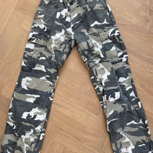 Camouflage cargopants från Mil-Tec XS - Säljer ett par grå, svarta och beige cargopants från Mil-Tec i storlek XS. Byxorna har klassiskt camouflage-mönster, flera stora fickor på sidorna och snörning vid bensluten. Materialet är slitstark bomull med syntet, perfekt för dig som gillar streetstyle.