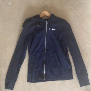 Mörkblå Nike hoodie med dragkedja - Säljer en mörkblå hoodie från Nike i storlek M. Tröjan har hel dragkedja, huva med snörning och två fickor framtill. Nike-loggan är broderad i vitt på bröstet. Perfekt för chill eller träning, riktigt skön och stilren design.