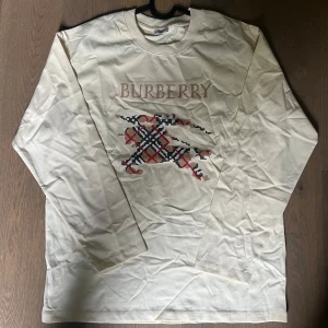 Burberry tröja  - Snygg långärmad tröja från Burberry i ljus beige bomull. Framsidan har ett stort broderat Burberry-tryck och klassiskt rutig mönster i rött, svart och beige. Perfekt för dig som gillar stilrena och exklusiva plagg med en twist. Knappt använd säljes pga av att det bara sitter i garderob… Storlek M