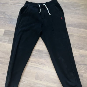 Svarta mjukisbyxor Polo Ralph Lauren - Svarta mjukisbyxor från Polo Ralph Lauren med vit snörning i midjan och röd broderad logga på ena höften. Byxorna har ribbade muddar vid bensluten och är tillverkade i mjukt bomullsmaterial. Perfekta för chill dagar eller när du vill vara bekväm.