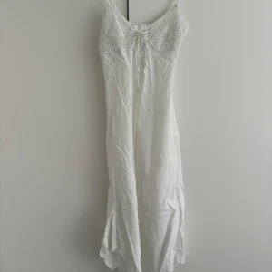 Vit långklänning Vintage - Supersöt vit långklänning med smala axelband och virkade detaljer upptill. Klänningen har snörning vid bysten och är gjord i mjukt linne. Perfekt för varma sommardagar och har en flowy siluett som ger boho-vibes. Vintage. Litet blått märke på insidan av bysten, men det syns ej på.