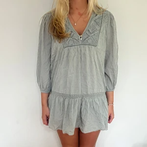 Ljusgrå kort klänning med broderi - Superfin ljusgrå kort klänning med v-ringning och broderade detaljer upptill. Klänningen har trekvartsärmar, små prickar i tyget och ett volangformat avslut nedtill.