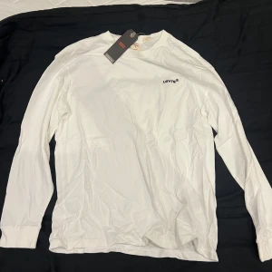 Vit långärmad tröja från Levi's - Säljer en stilren vit långärmad tröja från Levi's i mjuk bomull. Tröjan har rund hals och en liten svart Levi's-logga på bröstet. HELT NY MED LAPP KVAR