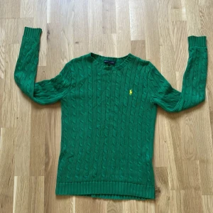 Grön kabelstickad tröja Ralph Lauren - Snygg grön kabelstickad tröja från Ralph Lauren med gul broderad logga på bröstet. Tröjan har rund hals, ribbade muddar och lång ärm. Perfekt för dig som gillar klassisk stil med en färgklick. Materialet är mjuk bomull som känns skönt mot huden.