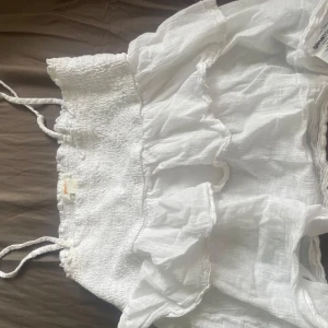 Vit volangkjol från Gina Tricot - Supersöt vit kjol från Gina Tricot med volanger och smock upptill. Kjolen är kort och har en luftig känsla, perfekt för varma dagar. Tillverkad i mjukt bomullstyg som känns skönt mot huden. Volangdetaljerna ger en extra feminin touch.