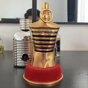 Jean Paul Gaultier Le Male le elixir - Exklusiv parfymflaska i form av en manlig torso, helblank i guld med horisontella ränder och en stor ring på toppen. Flaskan står på en rund röd sammetsbas som ger lyxig känsla. Perfekt för dig som gillar statement-dofter och snygg design.