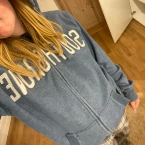 Hoodie med dragkedja  - Mysig blå hoodie från Reserved i storlek XL. Tröjan har dragkedja framtill, huva och stora fickor. Broderad vit text på bröstet ger en sportig vibe. Perfekt för chill dagar eller när du vill vara extra bekväm. Varan har tecken på noppror 