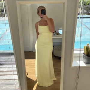 Ljusgul långklänning - Säljer en ljusgul långklänning med smala axelband och snygg öppen rygg med korsade band och knyt. Klänningen har en tight passform upptill och faller mjukt ner mot golvet. 