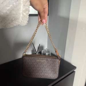 Brun axelväska från Michael Kors - Snygg brun axelväska från Michael Kors med kedjehandtag i guld och klassiskt MK-mönster. Väskan har dragkedja upptill och flera fack inuti för kort och småsaker. Perfekt storlek för det viktigaste och en stilren detalj till din outfit.