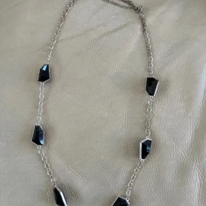 Silverhalsband med svarta stenar - Kedjehalsband i silverfärg med flera oregelbundna, svarta stenar i geometrisk form. Stenarna är infattade i metall och sitter utspridda längs hela halsbandet. Perfekt för dig som gillar en stilren men edgy look.