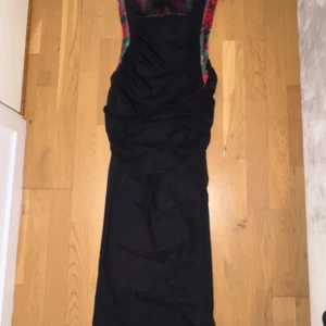 Svart långklänning Label Nue S - Supersnygg svart långklänning från Label Nue i storlek S. Klänningen har smala axelband och coola blommiga detaljer i rött och grönt längs sidorna och upptill. Den är figurnära med rynkade partier och dragkedja i ryggen. Perfekt för dig som vill sticka ut med en unik look!