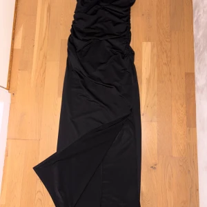 Svart långklänning med slits - Säljer en svart långklänning med snygga rynkade detaljer och en hög slits på sidan. Klänningen är axelbandslös och har en tight passform som framhäver figuren. Perfekt för dig som vill ha en stilren och elegant look.