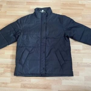 Ny svart pufferjacka i polyester - Svart pufferjacka med hög krage och raka linjer. Jackan har två snedställda fickor framtill och stängs med knappar. Tillverkad i polyester, perfekt för kyliga dagar. Enkel och stilren design som funkar till det mesta. Bröst 120cm, längd 73cm.