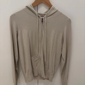 Beige hoodie med dragkedja från 157 - Snygg beige hoodie från 157 i storlek XS. Tröjan har huva med snörning, dragkedja framtill och ribbade muddar vid ärmslut och nederkant. Perfekt för chill dagar och enkel att matcha med jeans eller leggings.