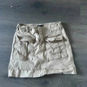 Beige kortkjol från gina tricot - Säljer en beige kortkjol från gina tricot i bomull. Kjolen har två stora fickor framtill med lock och bälteshällor i midjan. Storlek 38
