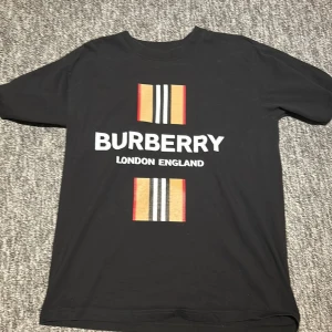 Svart Burberry t-shirt med tryck - Svart t-shirt från Burberry med klassiskt tryck framtill i vitt, beige, rött och svart. Stor logga och texten 'London England' på bröstet samt två block med Burberrys signaturränder. Tillverkad i 100% bomull och har korta ärmar.
