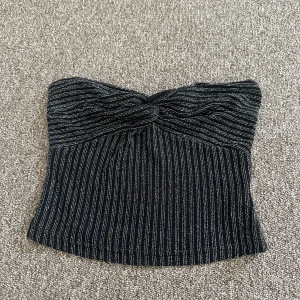 Glittrig svart tubtopp från SHEIN - Säljer en svart tubtopp från SHEIN i storlek XS. Toppen har smala silvriga ränder och glittrig finish, samt en snygg twistdetalj framtill. Den är endast använd fåtal gånger så det är jättebra skick på den. 