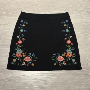 Svart kortkjol med broderade blommor - Svart kortkjol med färgglada broderade blommor längs sidorna. Kjolen har en rak och enkel form.