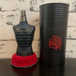 Jean Paul Gaultier Ultra Male parfym - Jean Paul Gaultier Ultra Male parfym i en ikonisk svart flaska formad som en manlig torso med mattsvarta och blanka ränder. Kommer med en svart metallcylinder med röd text och detaljer. Flaskan har metallförslutning och står på en röd sammetsliknande bas.