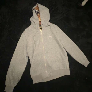 Grå hoodie från Burberry med dragkedja - Säljer en grå hoodie från Burberry med klassiskt rutigt foder i huvan. Tröjan har dragkedja framtill, snörning vid huvan och ribbade muddar. Perfekt för dig som gillar stilrena märkesplagg med en twist. Ikonisk Burberry-logga broderad på bröstet. Jätte fin Burberry kofta sällan använd 9/10 på skick. Ny pris~4000kr mitt pris är 800kr har du några frågor är det bara att höra av sig🤩