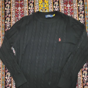 Svart kabelstickad tröja Polo Ralph Lauren - Kabelstickad svart tröja från Polo Ralph Lauren med klassisk röd broderad logga på bröstet. Tröjan har rund halsringning, långa ärmar och ribbade muddar. Perfekt för dig som gillar stilren och tidlös design.