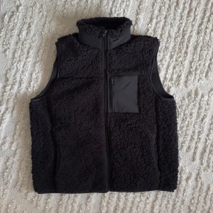 Fleeceväst - Säljer en oanvänd väst med dragkedja och en praktisk bröstficka. Perfekt för kyliga dagar och ger en mysig känsla. Västen har en hög krage för extra värme.