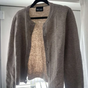 Beige kofta från Pieces - Säljer en mysig beige kofta från Pieces. Den har en klassisk design med knappar framtill och rund halsringning. Perfekt för att hålla sig varm under kyligare dagar.