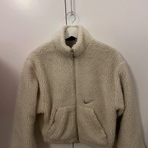 Beige teddyjacka från Nike - Mysig beige teddyjacka från Nike med dragkedja och hög krage. Jackan har två framfickor och en stor Nike-logga på ryggen. Perfekt för kyliga dagar.