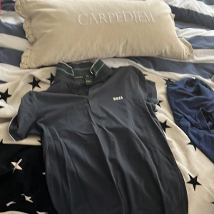 Svart pikétröja från Boss - Snygg svart pikétröja från Boss med gröna detaljer på kragen. Tröjan har en klassisk pikédesign med korta ärmar och knappar framtill. Perfekt för en stilren look.