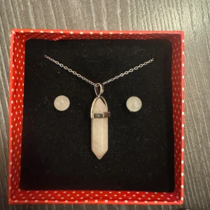 Halsband och örhängen med klar kvarts - Säljer ett set med halsband och örhängen i klar kvarts från The Mindful Frog. Halsbandet har en silverkedja med en spetsformad kvartssten. Örhängena har sterling silverstift och matchar halsbandet perfekt. Perfekt för att ge en energigivande touch till din stil.