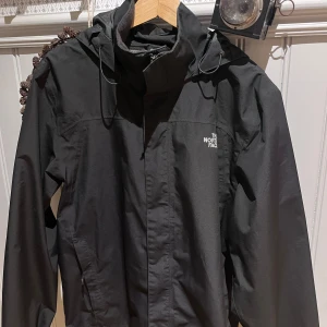 Svart vindjacka från The North Face - Säljer en stilren svart vindjacka från The North Face. Jackan har en praktisk huva och dragkedja framtill. Perfekt för blåsiga dagar och håller dig skyddad mot väder och vind. Modellen är runt 180 och väger 75kg.