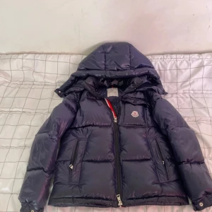 Mörkblå dunjacka från Moncler - Säljer en snygg mörkblå dunjacka från Moncler med huva och dragkedja. Jackan har en glansig finish och är perfekt för kyliga dagar. Den har långa ärmar och en klassisk Moncler-logga på bröstet.