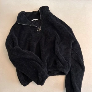 Svart teddy tröja från Chiquelle - Mysig svart fleece hoodie med en halv dragkedja och en rund dragkedjering. Perfekt för kyliga dagar när du vill hålla dig varm och bekväm. Den har en avslappnad passform och långa ärmar. Något croppad