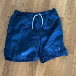Blå badshorts från Polo Ralph Lauren - Snygga blå badshorts från Polo Ralph Lauren med vit snörning i midjan. Perfekta för sommarens badäventyr. Den klassiska loggan i rött ger en stilren touch.