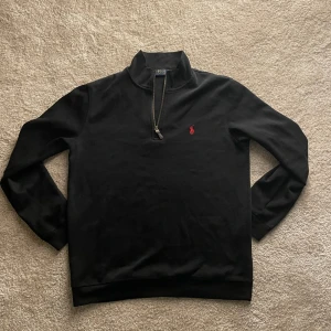 Svart tröja med dragkedja från Polo Ralph Lauren - Säljer en stilren svart tröja från Polo Ralph Lauren med dragkedja och röd logga på bröstet. Perfekt för en avslappnad men snygg look. Tröjan har långa ärmar och en hög krage.