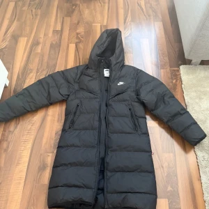 Svart lång dunjacka  Nike - Säljer en svart lång dunjacka från Nike med huva och dragkedja. Jackan har en stilren design med Nike-logga på bröstet och är perfekt för kalla dagar. Den är utrustad med Storm-FIT-teknologi för extra skydd mot väder och vind.