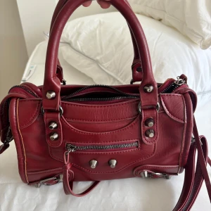 Röd väska balenciaga liknande - Snygg röd skinnväska med både handtag och axelrem. Den har även ett kortfack. En liten ficka på utsidan och insidan. Fint skick! Pris kan diskuteras vid snabb affär. 