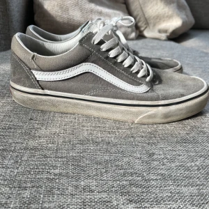 Grå sneakers från Vans - Snygga grå sneakers från Vans med vit sula och klassisk sidostrip. Skorna har snörning och en bekväm passform, perfekt för vardagsbruk. Yttersulan är i gummi med det ikoniska rutmönstret för bra grepp.