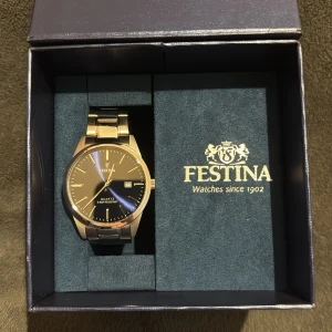 Silverfärgad klocka från Festina - Snygg silverfärgad klocka från Festina med marinblå urtavla och datumvisning. Klockan har ett klassiskt armband i metall och levereras i en elegant blå förpackning.