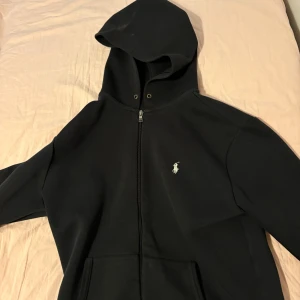Svart hoodie från Ralph Lauren - Säljer en stilren svart hoodie från Ralph Lauren med dragkedja och det klassiska logotypbroderiet på bröstet. Tröjan har långa ärmar och en praktisk huva. Perfekt för en avslappnad look.