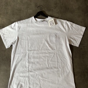 Vit t-shirt från Loewe - Säljer en stilren vit t-shirt från Loewe med ett diskret broderat logotypmärke på bröstet. T-shirten har en klassisk rund hals och korta ärmar. Perfekt för en minimalistisk look.