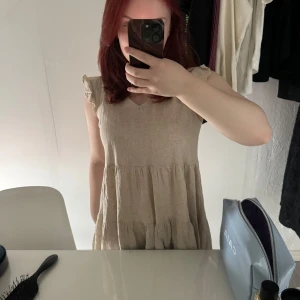 Beige klänning - Säljer en söt beige klänning från Shein med volangdetaljer vid ärmarna. Klänningen har en v-ringad hals och är ärmlös, perfekt för varma dagar. Tillverkad i ett lätt och luftigt material.