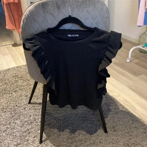 Svart topp med volangdetaljer från Zara - Säljer en snygg svart topp från Zara med volangdetaljer längs ärmarna. Perfekt för att ge en extra touch till din outfit. Toppen har en rund halsringning och är tillverkad i ett mjukt material.💓