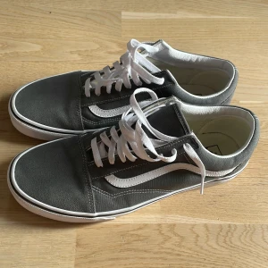 Grå sneakers från Vans - Snygga grå sneakers från Vans med vit sula och klassisk vit sidostrimma. Skorna har snörning och en rund tå, perfekt för en avslappnad stil. Gummisulan ger bra grepp och komfort.